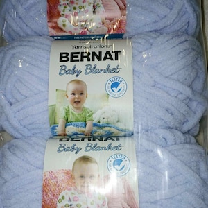 May include: Two skeins of light blue Bernat Baby Blanket yarn. The yarn is packaged in a clear plastic bag with a label that features a baby wearing a pink and white patterned onesie. The label also includes the text "Yarnspirations" and "Bernat Baby Blanket".