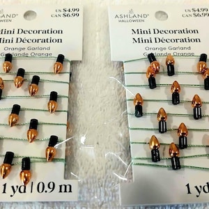 May include: Two packages of Ashland Halloween mini orange garland, each measuring 1 yard. The garland features small, metallic orange light bulb-shaped charms with black bases, strung on thin green thread on white cardboard backing.