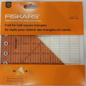 May include: An orange and clear Fiskars folding square ruler for quilting. The ruler is marked with measurements and angles, and the packaging includes instructions for folding half-square triangles. The ruler is 8.375 inches when opened.