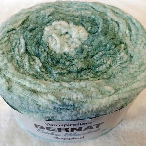 May include: A round ball of Bernat Baby Blanket dappled yarn in shades of teal and white. The yarn is soft and plush, ideal for baby blankets and other cozy projects. The label reads "Bernat Baby Blanket dappled".