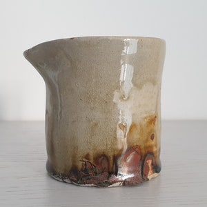 May include: A small, off-white ceramic jug with a brown glaze. The jug has a slightly irregular shape and a rounded spout.