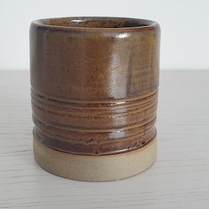 Puede incluir: Una taza de cerámica marrón con una base marrón claro. La taza tiene un acabado esmaltado y una forma ligeramente redondeada.