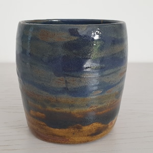 Puede incluir: Una taza de cerámica con un esmalte azul, marrón y dorado. La taza tiene una forma redondeada y un acabado liso y brillante.
