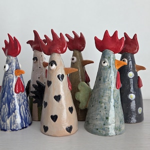 Może przedstawiać: Kolekcja ceramicznych figurek kogutów z czerwonymi grzebieniami, pomarańczowymi dziobami i różnymi wzorami. Koguty są w kształcie stożka, niektóre z motywami serca, inne z niebieskim, zielonym i czarnym wykończeniem. Ustawione na białej powierzchni.