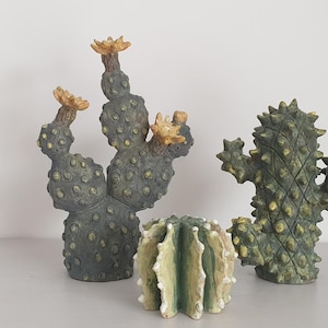Può includere: Tre sculture di cactus in ceramica in tonalità verdi e marroni. Il cactus a sinistra ha fiori gialli e una superficie testurizzata. Il cactus a destra ha un motivo a forma di diamante spinoso. Il cactus al centro è rotondo e ha un motivo a strisce.