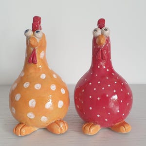 Könnte beinhalten: Zwei skurrile Keramik-Hühnerfiguren. Eine ist orange mit weißen Punkten, die andere rot mit weißen Punkten. Beide haben übertriebene Merkmale, darunter große Augen, rote Kämme und orangefarbene Füße.