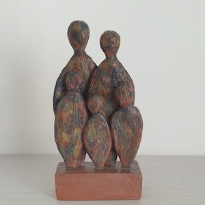 Może przedstawiać: Ceramiczna rzeźba przedstawiająca rodzinę, z czterema postaciami stojącymi razem. Postacie są stylizowane i abstrakcyjne, w stonowanych odcieniach brązu, błękitu i zieleni. Rzeźba stoi na prostokątnym cokole.