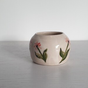 Peut inclure: Un petit pot en céramique blanc de forme arrondie avec un motif floral. Le pot est décoré de fleurs roses et vertes peintes sur le côté.