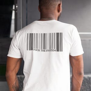 May include: White t-shirt with a black barcode design and the text "FUCK THE SYSTEM" printed on the back. The shirt is a crew neck style and appears to be made of a soft material.
