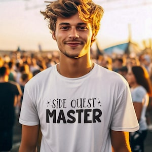 May include: White t-shirt with the text "SIDE QUEST MASTER" in black. The shirt has a crew neck and short sleeves. The person wearing the shirt is smiling, with a crowd in the background.