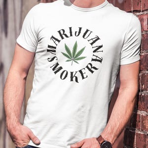 May include: White t-shirt with a circular design featuring the text "MARIJUANA SMOKER" around a green cannabis leaf. The shirt is plain and has a crew neck.