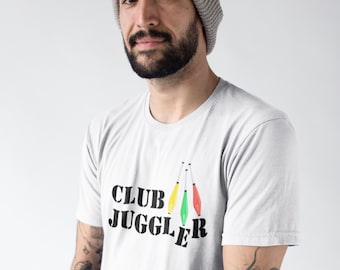 May include: A white t-shirt with the text "CLUB JUGGLER" in black, with three juggling clubs in yellow, green, and red. The person in the image is wearing a gray beanie.