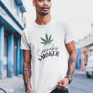May include: A light gray t-shirt featuring a green cannabis leaf and the words "Cannabis SMOKER" in black script. The shirt is worn by a person with tattoos and a watch.