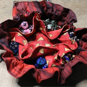 May include: Red fabric dice bag with a heart pattern and a drawstring closure. The bag is filled with various colored dice, including blue, pink, and green.