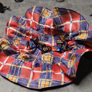 May include: A red, blue, and yellow plaid fabric dice bag with a button closure. The bag is filled with various colored dice.