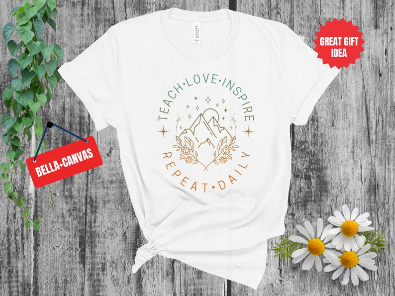 May include: White t-shirt with a green, brown, and orange graphic design that says "Teach Love Inspire Repeat Daily" with a mountain and stars in the center.