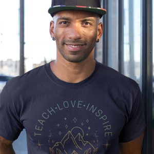 May include: A man wearing a black baseball cap with a red, white, and blue stripe, and a navy blue t-shirt with a graphic design of mountains, stars, and the text "Teach Love Inspire Repeat Daily".