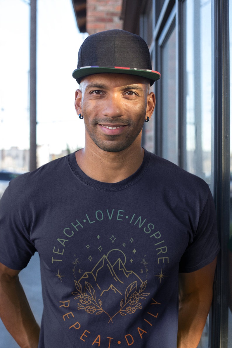 May include: A man wearing a black baseball cap with a red, white, and blue stripe, and a navy blue t-shirt with a graphic design of mountains, stars, and the text "Teach Love Inspire Repeat Daily".