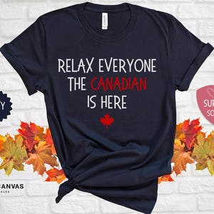 以下が含まれることがあります： 紺色のTシャツに、「リラックスみんなカナダ人はここにいる」という白い文字が書かれています。テキストの下には赤いカエデの葉があります。