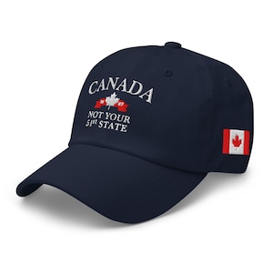 May include: A navy blue baseball cap with white embroidery that reads "CANADA NOT YOUR 51st STATE" with a red and white Canadian flag on the side. The embroidery also includes a red maple leaf with the years "1867" above and below it.