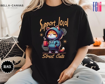 May include: Black t-shirt with a cartoon cat wearing a blue hoodie and holding a cup of coffee. The text "Support Local Street Cats" is printed on the shirt.