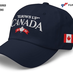 May include: A navy blue baseball cap with white embroidered text that reads "Elbows Up" Canada 1867. The cap has a red Canadian flag patch on the side.