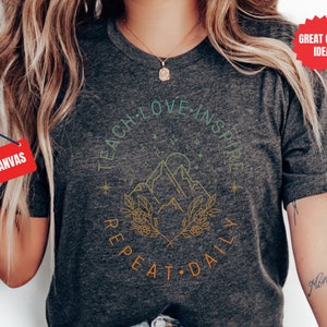 May include: A heather grey t-shirt with a graphic design featuring a mountain range, a sun, and stars. The text "Teach Love Inspire Repeat Daily" is written in a circle around the graphic.
