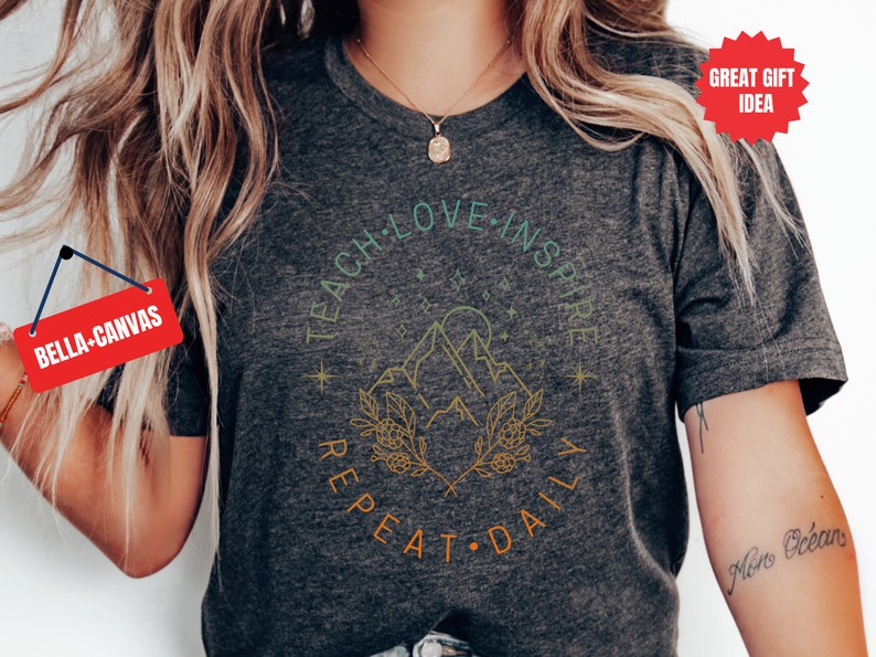 May include: A heather grey t-shirt with a graphic design featuring a mountain range, a sun, and stars. The text "Teach Love Inspire Repeat Daily" is written in a circle around the graphic.