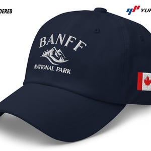 Pode incluir: Um boné de beisebol azul marinho com texto bordado em branco que diz "Banff National Park" e um gráfico de montanha branco. O boné também tem um pequeno patch de bandeira canadense vermelha e branca no lado.