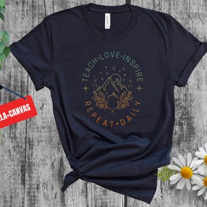 May include: A navy blue t-shirt with a graphic design of mountains, stars, and foliage. The text "Teach Love Inspire Repeat Daily" is written in a circle around the graphic.