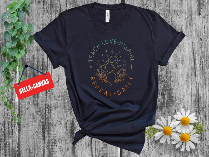 May include: A navy blue t-shirt with a graphic design of mountains, stars, and foliage. The text "Teach Love Inspire Repeat Daily" is written in a circle around the graphic.