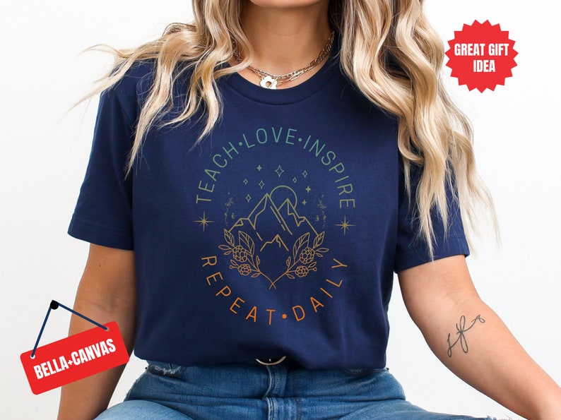 May include: Navy blue t-shirt with a graphic design of mountains, stars, and leaves. The text "Teach Love Inspire Repeat Daily" is printed in a circle around the graphic.