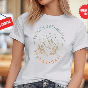 May include: White t-shirt with a graphic design of mountains, stars, and foliage. The text "Teach Love Inspire Repeat Daily" is written in a circle around the graphic.