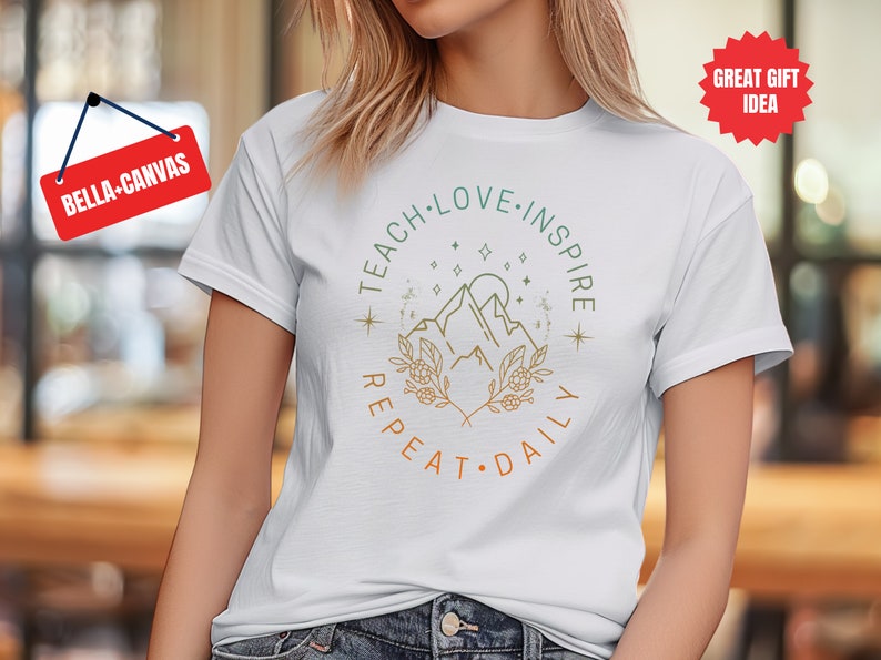 May include: White t-shirt with a graphic design of mountains, stars, and foliage. The text "Teach Love Inspire Repeat Daily" is written in a circle around the graphic.