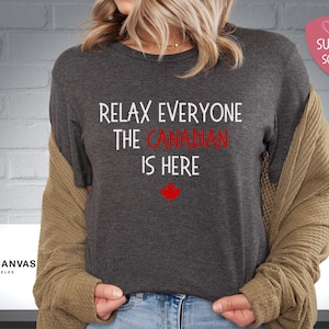 以下が含まれることがあります： 暗いグレーのTシャツに、「リラックスみんなカナダ人ここにいる」という白い文字が書かれています。テキストの下には赤いカエデの葉があります。