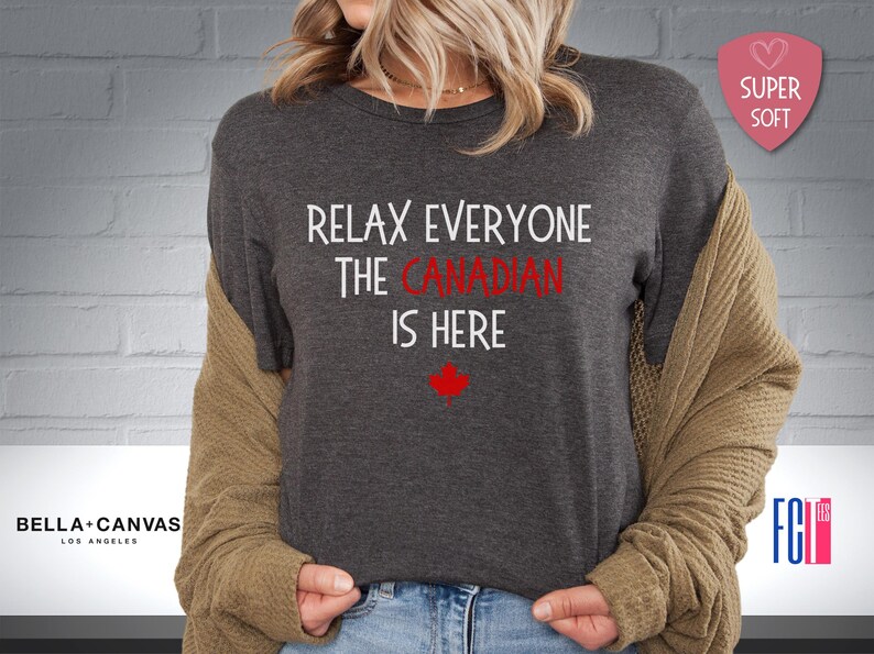 以下が含まれることがあります： 暗いグレーのTシャツに、「リラックスみんなカナダ人ここにいる」という白い文字が書かれています。テキストの下には赤いカエデの葉があります。