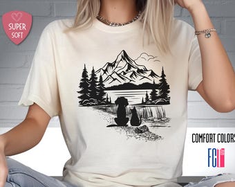 May include: A white t-shirt with a black silhouette design of a dog and cat sitting in front of a mountain landscape with trees and a lake. The text "COMFORT COLORS" is printed on the bottom right corner of the shirt.