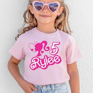 May include: A light pink t-shirt with a graphic featuring a silhouette, the number 5, and the name "Rylee" in a playful font. The shirt is worn by a young person wearing heart-shaped sunglasses.