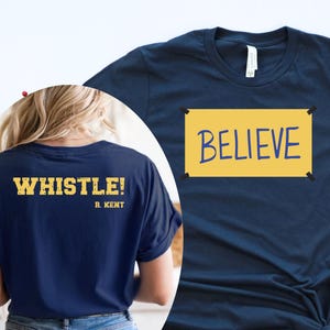 May include: Navy blue t-shirt with the word "BELIEVE" in blue on a yellow rectangle. The back of the shirt says "WHISTLE!" in gold lettering. The shirt is paired with blue jeans and white sneakers.