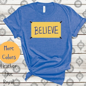 May include: A royal blue t-shirt with a yellow sign that says "BELIEVE" in black lettering. The sign has black tape on the corners.