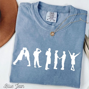 May include: A blue jean colored t-shirt with the word "ARMY" in white block letters. The letters are formed by white silhouettes of people. A gold necklace with a pendant is on the shirt. The shirt has a Comfort Colors tag.