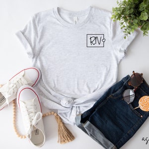 May include: A grey t-shirt with a black square and the text "RN" with a small heart inside the square. The t-shirt is on a white background with a pair of white trainers, a beaded necklace, a pair of denim shorts, and sunglasses.