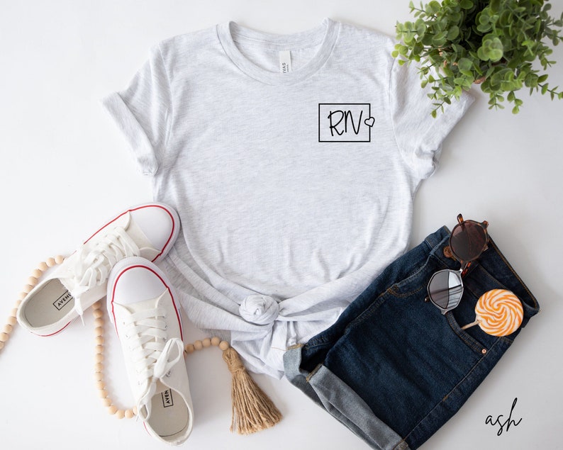 May include: A grey t-shirt with a black square and the text "RN" with a small heart inside the square. The t-shirt is on a white background with a pair of white trainers, a beaded necklace, a pair of denim shorts, and sunglasses.