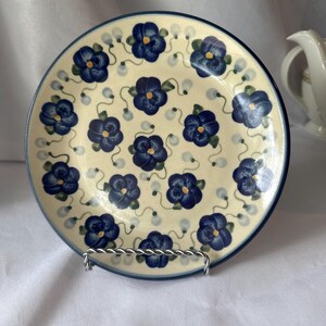 Op de afbeelding: Een ronde keramische plaat met een crèmekleurige achtergrond en een blauw bloemmotief. De plaat heeft een donkerblauwe rand en staat op een zilveren metalen standaard. Het bloemmotief omvat donkerblauwe bloemen en groene stelen.