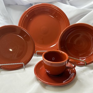 May include: A collection of orange-red ceramic tableware, featuring plates, bowls, a teacup, and a saucer. The items are displayed on a white surface, highlighting their glossy finish and traditional design. The set includes a large dinner plate, a smaller plate, and a bowl.