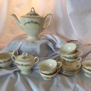 May include: A vintage cream-colored tea set with floral accents. The set includes a teapot, sugar bowl, creamer, cups, and saucers. The floral design features pink, yellow, and blue flowers with gold trim. The teapot stands approximately 8 inches tall.