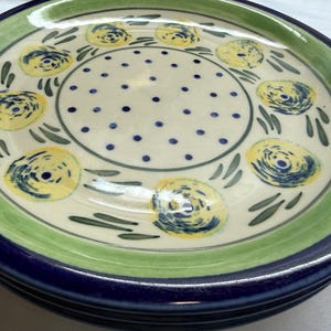 May include: Stack of ceramic plates with a floral design. The plates have a cream-colored center with blue dots and yellow floral patterns. The rims are green and blue, creating a layered effect. A decorative and functional addition to any kitchen.