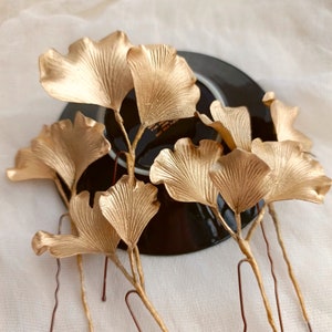 May include: Gold ginkgo leaf hair pins. The leaves are arranged in a cluster and are attached to thin gold wire stems. The pins are perfect for adding a touch of elegance to any hairstyle.