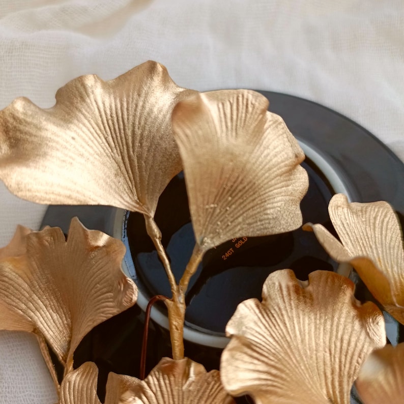 May include: A close-up of a golden ginkgo leaf arrangement on a black plate. The leaves are arranged in a fan shape and have a delicate, textured surface. The plate has a subtle sheen and a simple design.