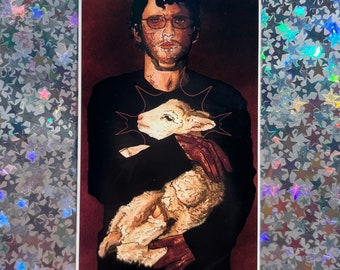 May include: A man with dark hair and glasses is holding a white lamb in his arms. He is wearing a black shirt and has a dark red background.